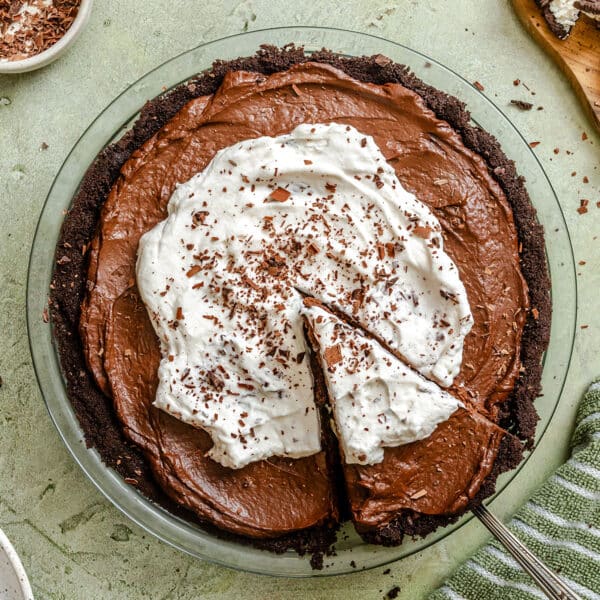 A chocolate avocado pie with a chocolate crust, topped with whipped cream and chocolate shavings, with one slice being cut and partially removed.