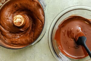 Chocolate mousse mixture in a food processor next to a bowl of melted chocolate with spatula on a light countertop.