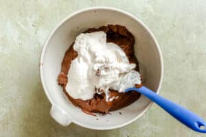 A mixing bowl with whipped cream being folded into melted chocolate using a blue spatula.