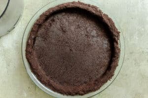 A glass pie dish filled with an even layer of chocolate cookie crumb crust, pressed on the bottom and up the sides.