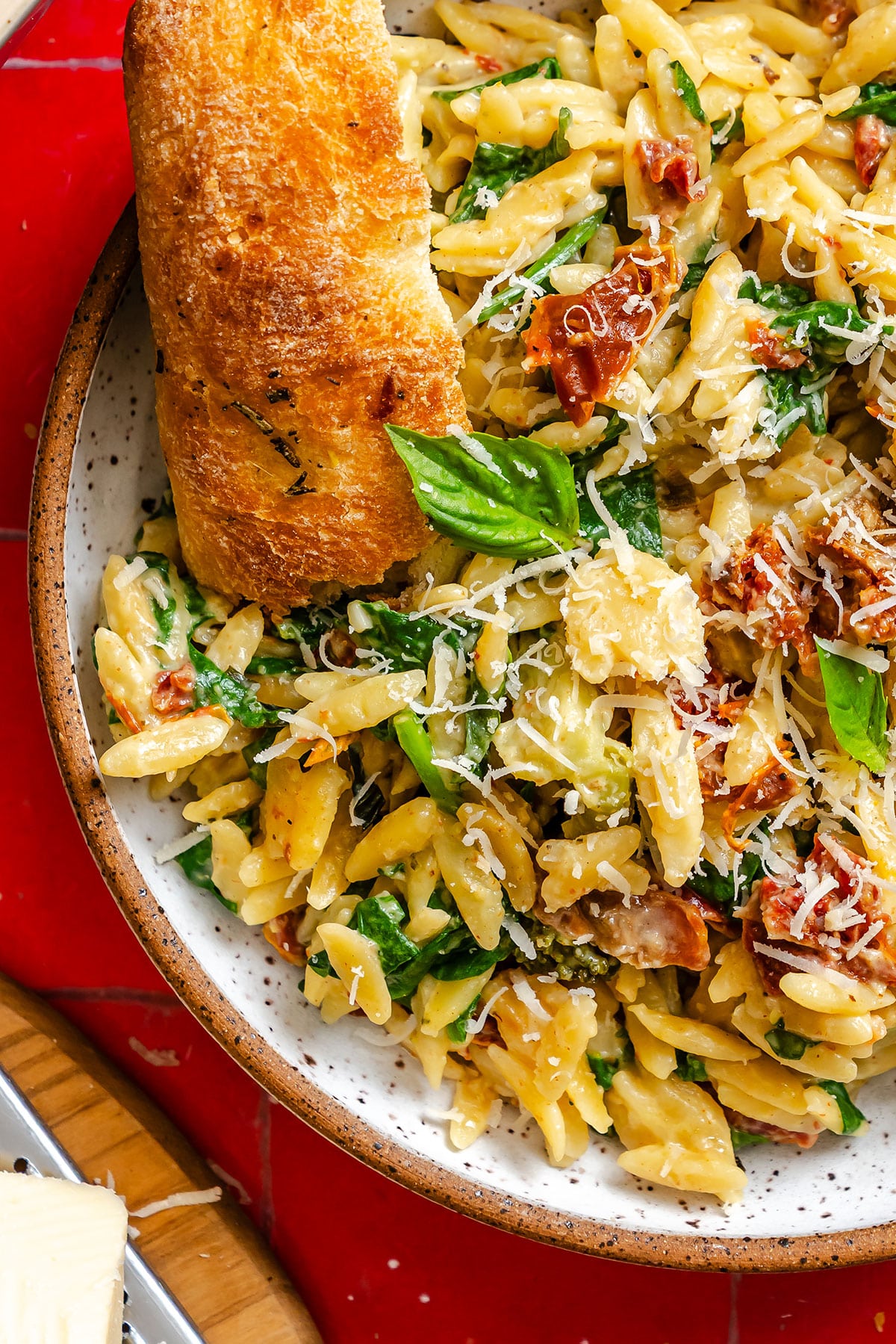 A plate of orzo pasta with spinach, sun-dried tomatoes, grated cheese, fresh basil, and a slice of toasted bread.