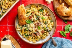 A plate of pasta with sun-dried tomatoes, spinach, grated cheese, and basil, served with a slice of bread on a red tiled surface.