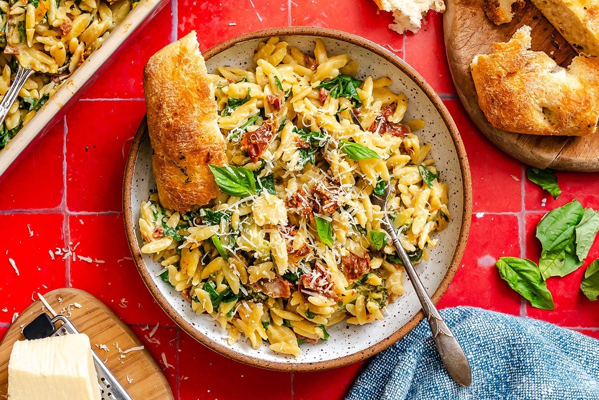 A plate of pasta with sun-dried tomatoes, spinach, grated cheese, and basil, served with a slice of bread on a red tiled surface.