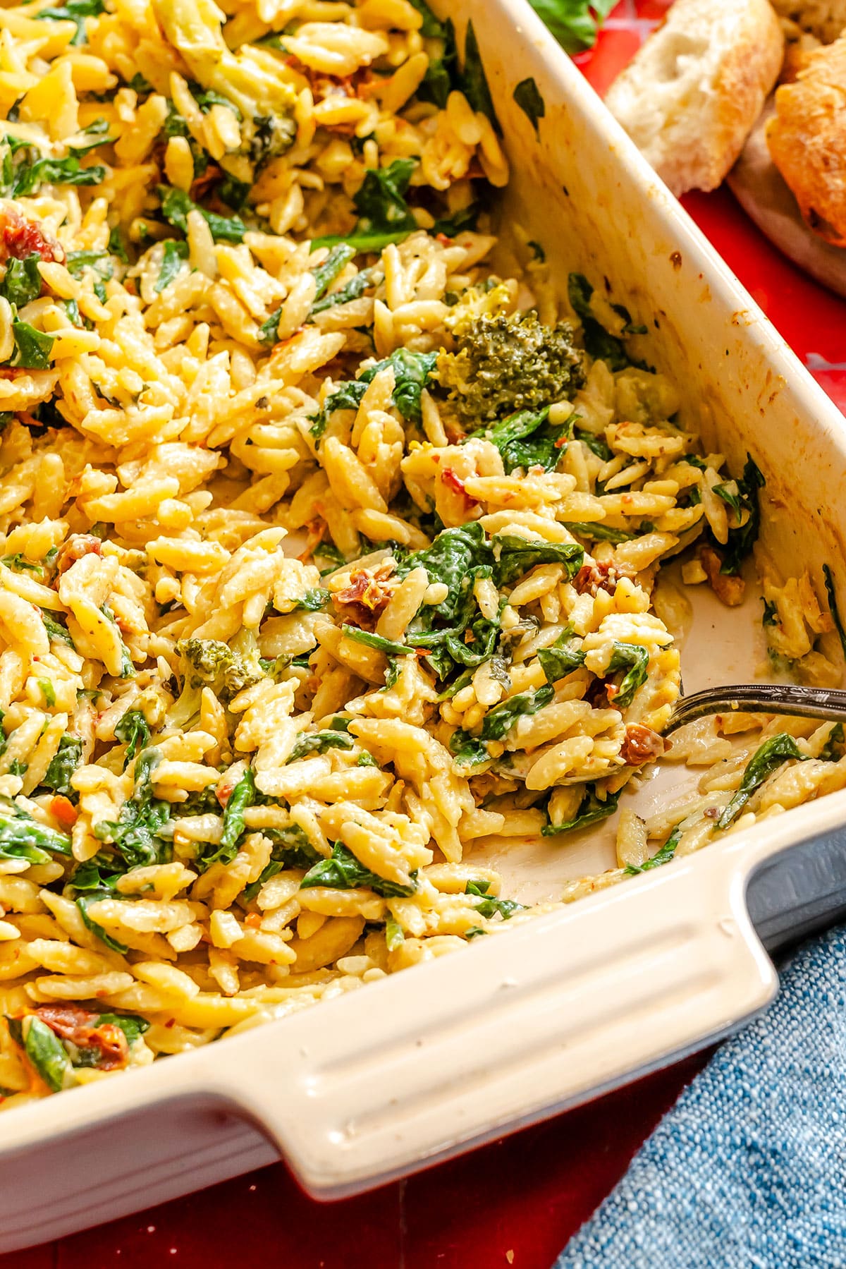 Baked orzo pasta mixed with spinach, sun-dried tomatoes, and pesto sauce in a rectangular casserole dish with a metal serving spoon.