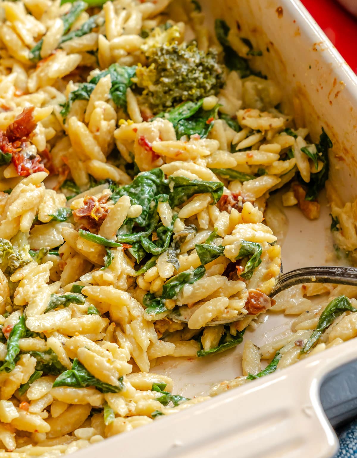 Baked orzo pasta dish with spinach, sun-dried tomatoes, broccoli, and melted cheese, partially served with a metal spoon in a casserole dish.