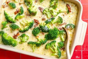 Casserole dish with broccoli florets and sun-dried tomatoes in a creamy sauce, with a wooden spoon resting inside.