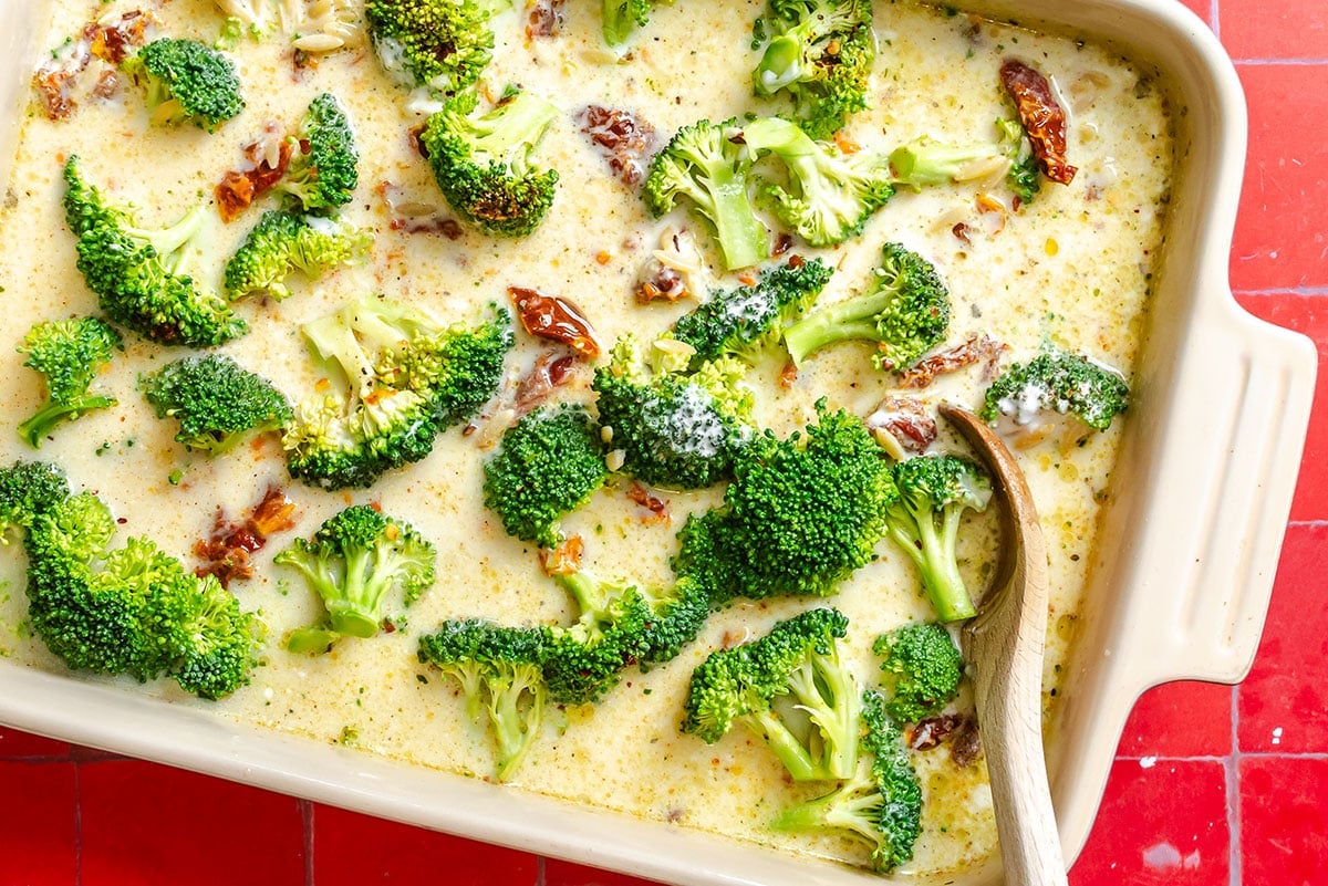 Casserole dish with broccoli florets and sun-dried tomatoes in a creamy sauce, with a wooden spoon resting inside.
