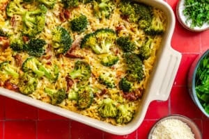 Baked casserole with pasta, broccoli florets, and a creamy sauce in a white dish, surrounded by bowls of chopped herbs, leafy greens, and grated cheese on a red tiled surface.