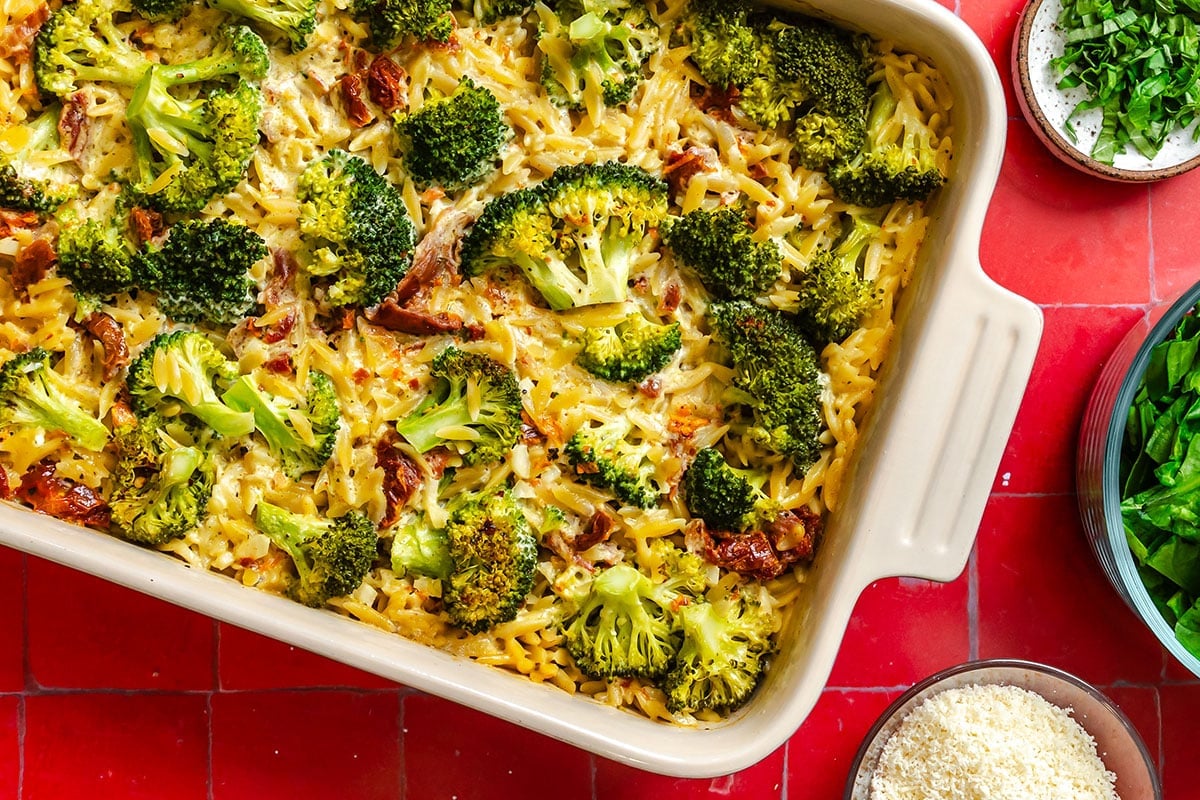 Baked casserole with pasta, broccoli florets, and a creamy sauce in a white dish, surrounded by bowls of chopped herbs, leafy greens, and grated cheese on a red tiled surface.