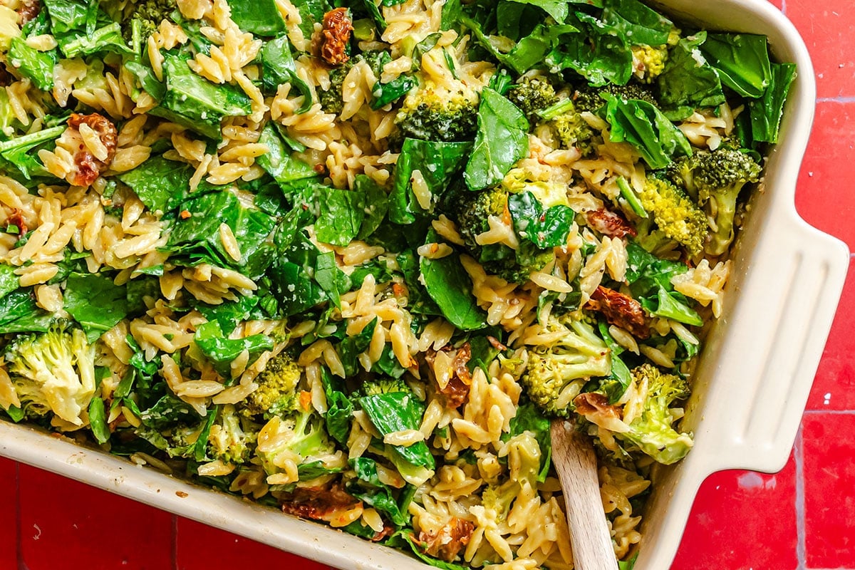 A casserole dish filled with orzo pasta, spinach, broccoli, and sun-dried tomatoes, stirred with a wooden spoon, on a red surface.