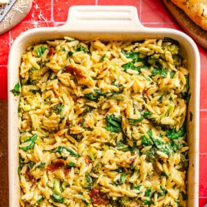 A baked dish filled with creamy marry me orzo pasta, spinach, broccoli, and sundried tomatoes on a red tile counter.