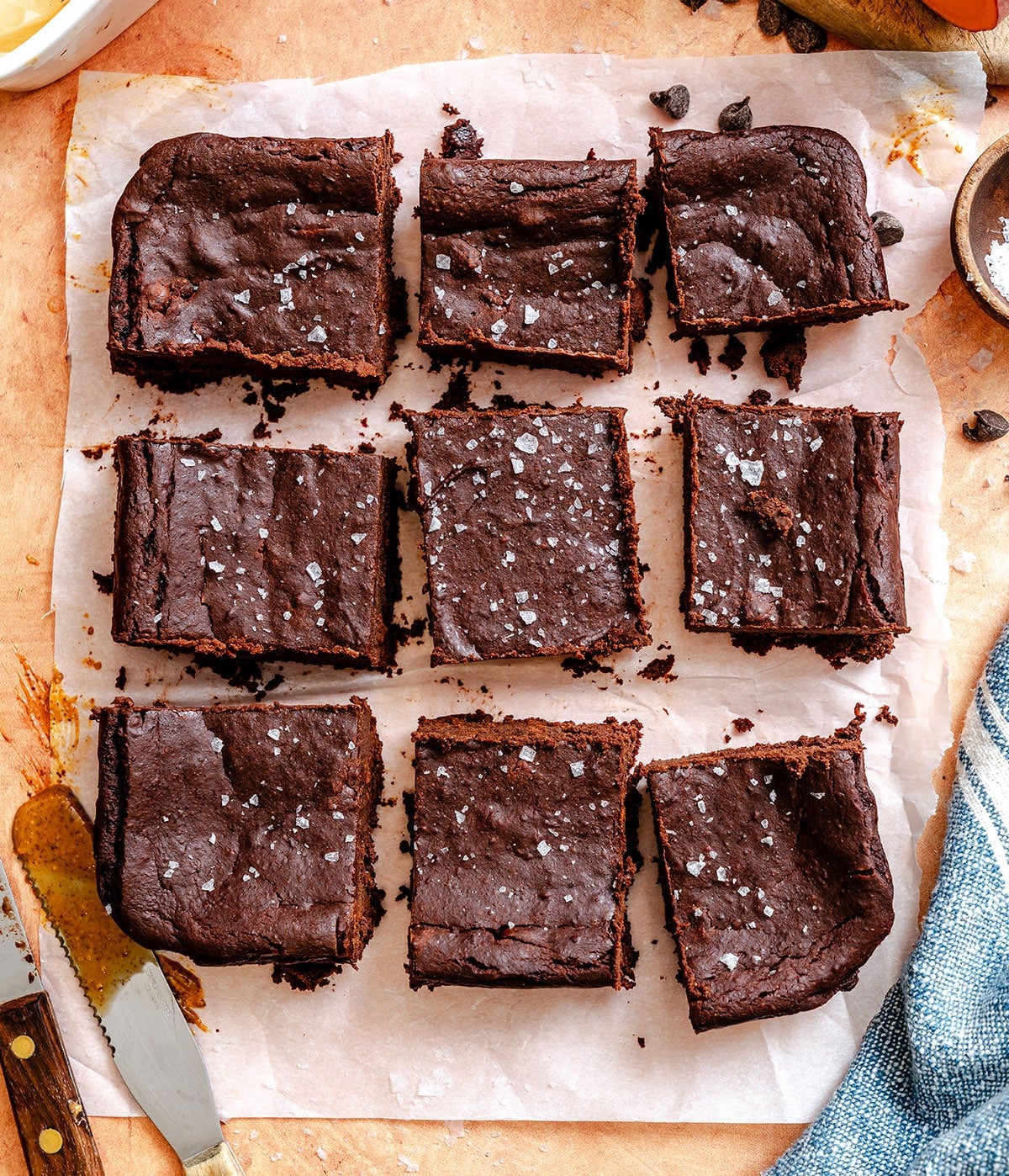 Nine chocolate brownies, each topped with coarse sea salt, are arranged in a 3x3 grid on parchment paper atop a wooden surface.