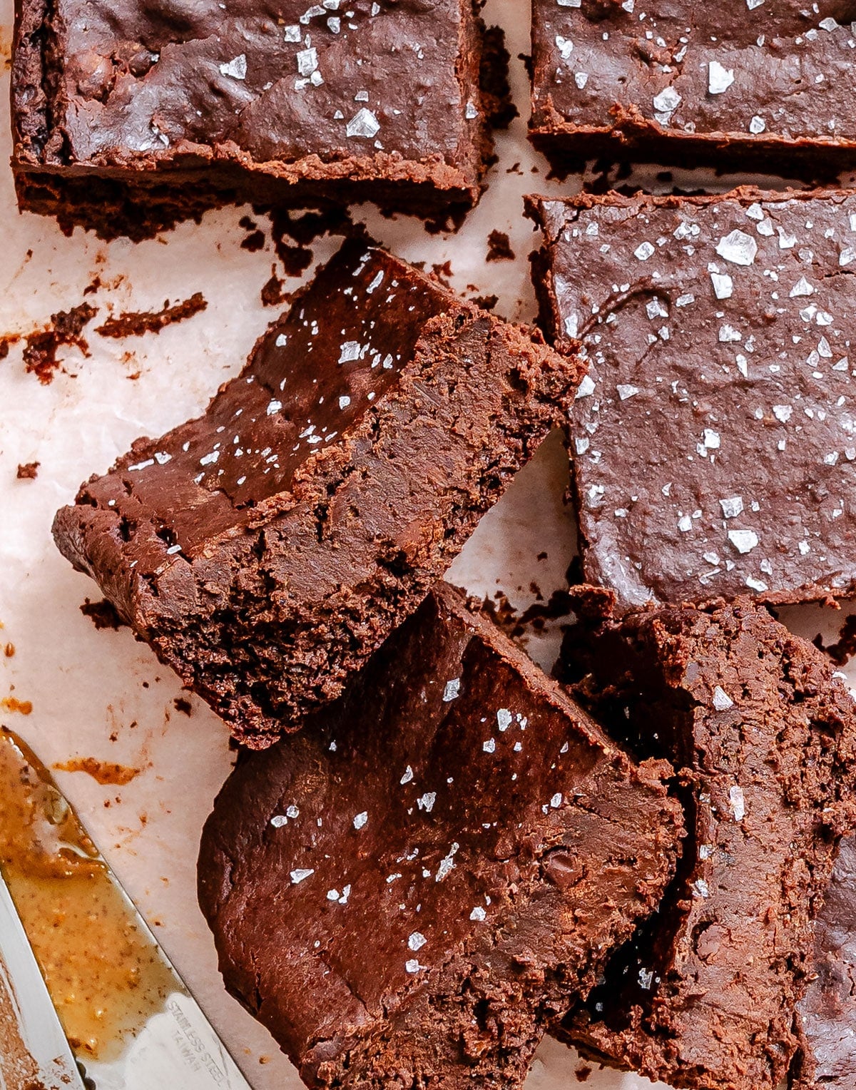 Several chocolate brownies topped with sea salt are arranged on parchment paper, with one piece turned to show a fudgy texture.