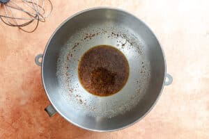A large metal mixing bowl with a small amount of dark liquid and spices at the bottom, placed on a light brown countertop. A metal whisk is visible in the top left corner.
