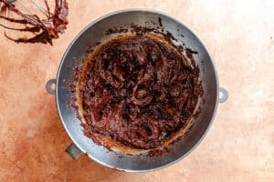 A metal mixing bowl filled with chocolate cake batter sits on a brown countertop, with a beater covered in batter nearby.