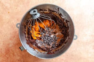 A metal mixing bowl with a whisk attachment contains chocolate batter, chocolate chips, pumpkin puree, and spices on a light brown surface.