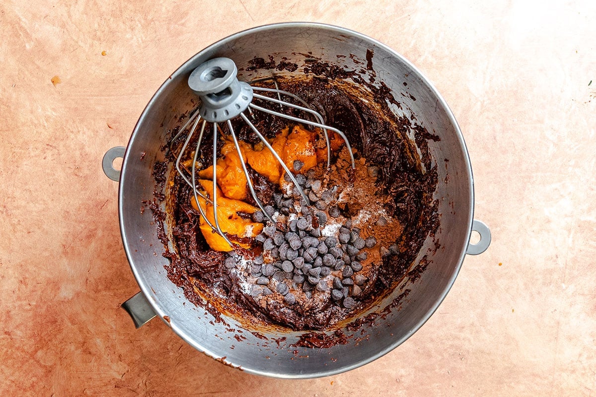 A metal mixing bowl with a whisk attachment contains chocolate batter, chocolate chips, pumpkin puree, and spices on a light brown surface.