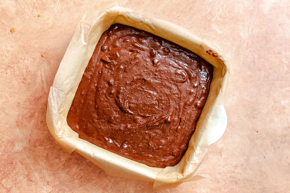 A square baking pan lined with parchment paper filled with chocolate brownie batter, photographed from above on a light brown surface.