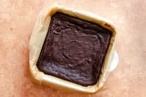 A baked pan of chocolate brownies sits on parchment paper in a square baking dish on a light brown surface.