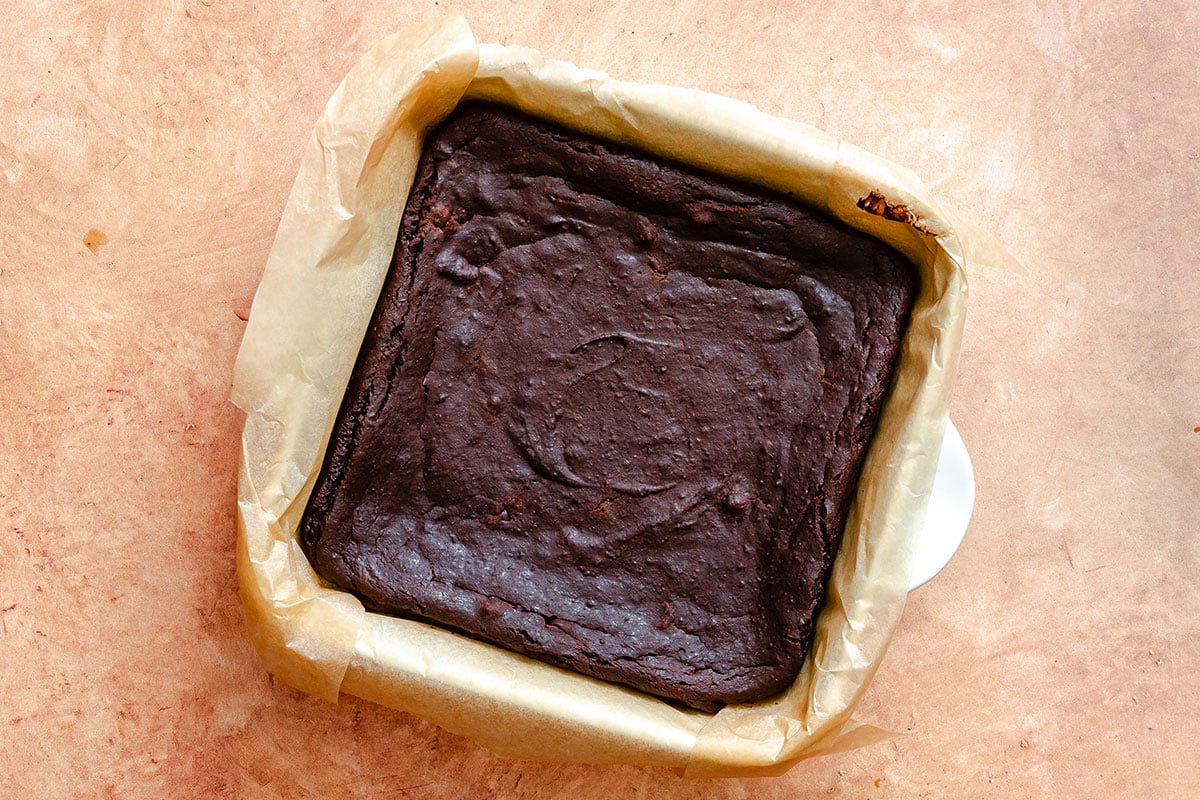 A baked pan of chocolate brownies sits on parchment paper in a square baking dish on a light brown surface.