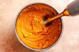 A bowl of orange sweet potato puree being blended with an immersion blender on a light brown surface.