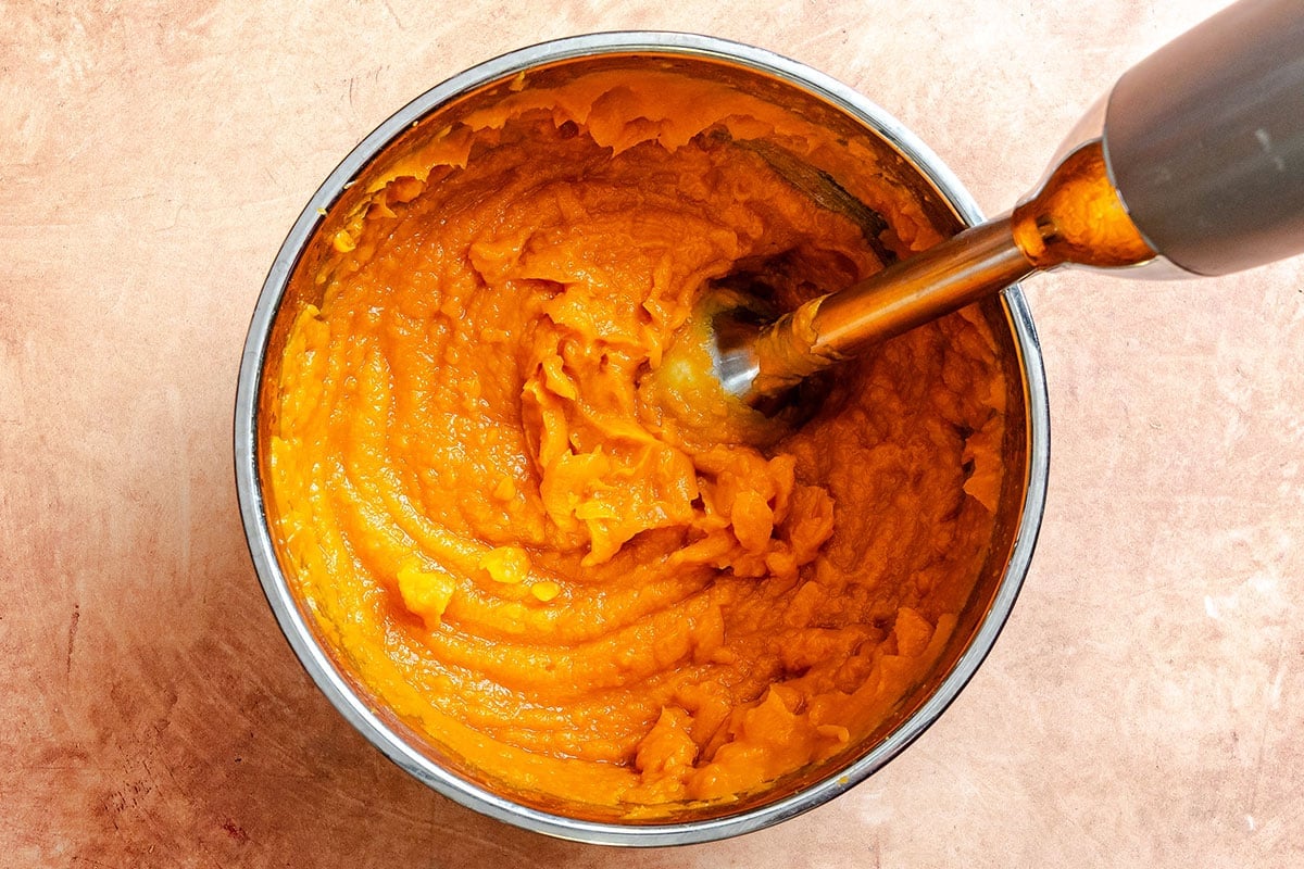 A bowl of orange sweet potato puree being blended with an immersion blender on a light brown surface.