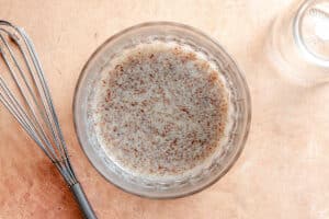 A glass bowl filled with a mixture of water and ground flaxseed sits on a light brown surface next to a metal whisk and an empty glass jar.