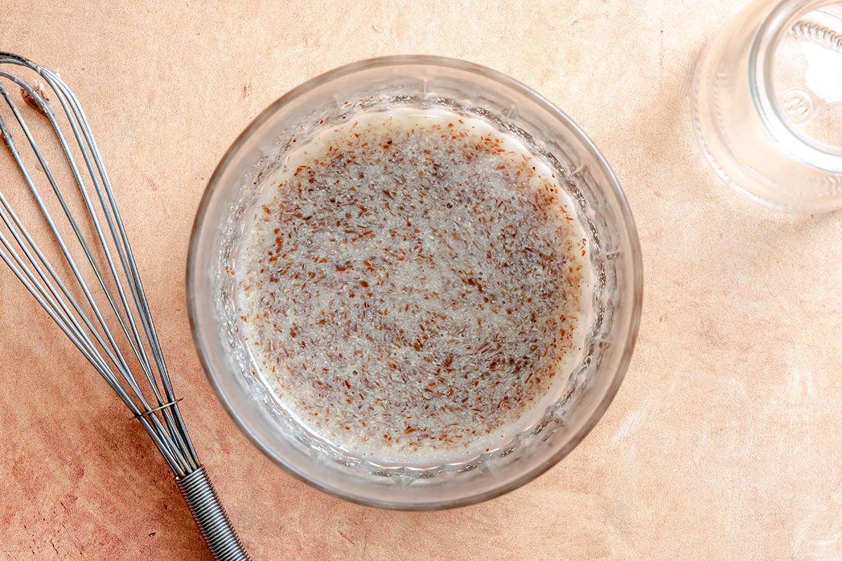 A glass bowl filled with a mixture of water and ground flaxseed sits on a light brown surface next to a metal whisk and an empty glass jar.
