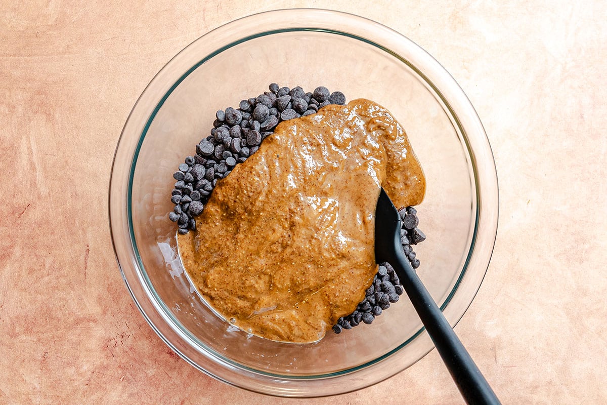 A glass bowl containing chocolate chips and nut butter being mixed with a black spatula, placed on a light brown surface.