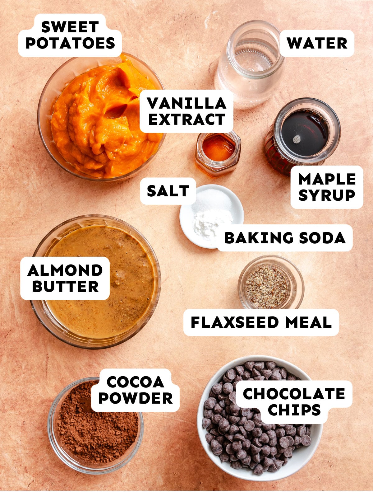 Top-down view of labeled ingredients in bowls and jars: sweet potatoes, almond butter, cocoa powder, vanilla extract, salt, baking soda, flaxseed meal, water, maple syrup, and chocolate chips.