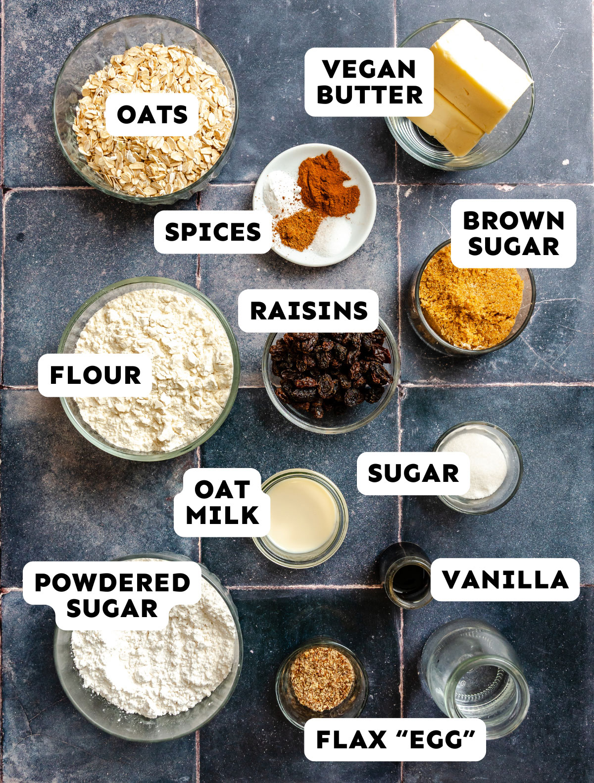 A flat lay of labeled baking ingredients including oats, vegan butter, brown sugar, flour, raisins, oat milk, sugar, powdered sugar, spices, vanilla, and flax “egg” on a tiled surface.