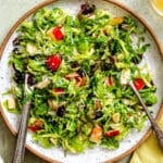 A plate of shredded Brussels sprouts salad with apple slices, dried cranberries, almonds, and shredded cheese, with two serving utensils.