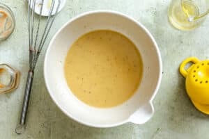 A mixing bowl filled with light brown liquid mixture sits on a countertop with a whisk, lemon squeezer, and various kitchen utensils nearby.