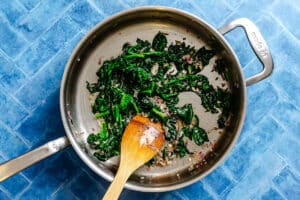 A stainless steel pan with sautéed spinach and onions, stirred with a wooden spatula, sits on a blue tiled surface.