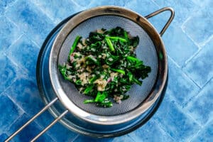 Chopped cooked spinach and onions draining in a fine mesh strainer over a metal bowl on a blue tiled surface.