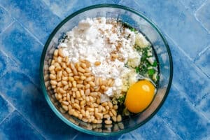A glass bowl containing pine nuts, ricotta cheese, a raw egg, seasonings, and chopped green herbs on a blue tiled surface.