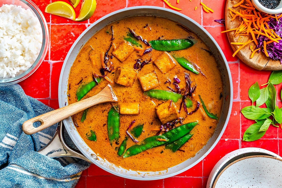 A skillet filled with tofu and vegetables in red curry sauce, surrounded by rice, lime wedges, shredded carrots, purple cabbage, and fresh basil on a red tiled surface.
