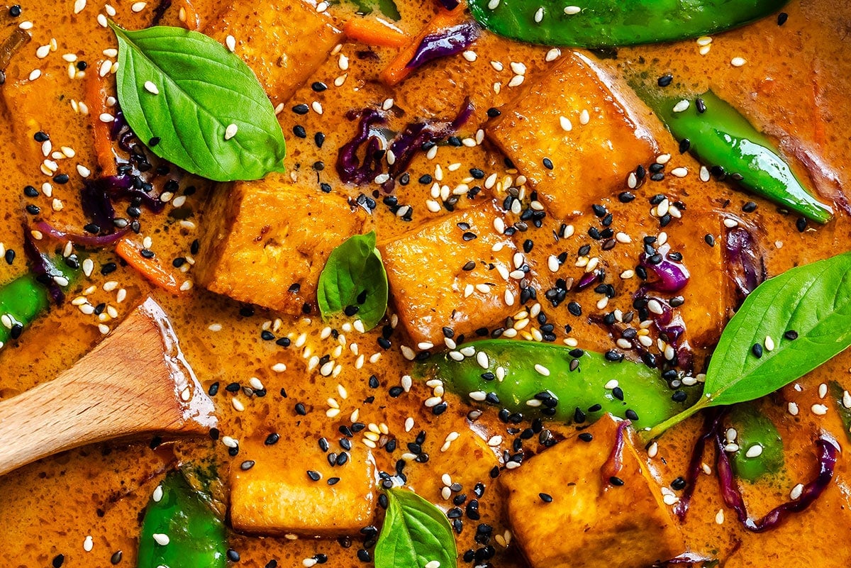 Close-up of tofu cubes, snap peas, and basil leaves in a creamy red curry sauce, garnished with black and white sesame seeds. A wooden spoon rests in the dish.
