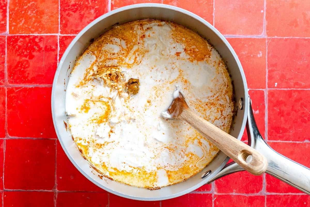 A saucepan with a creamy orange sauce being stirred with a wooden spoon, on a red tiled surface.