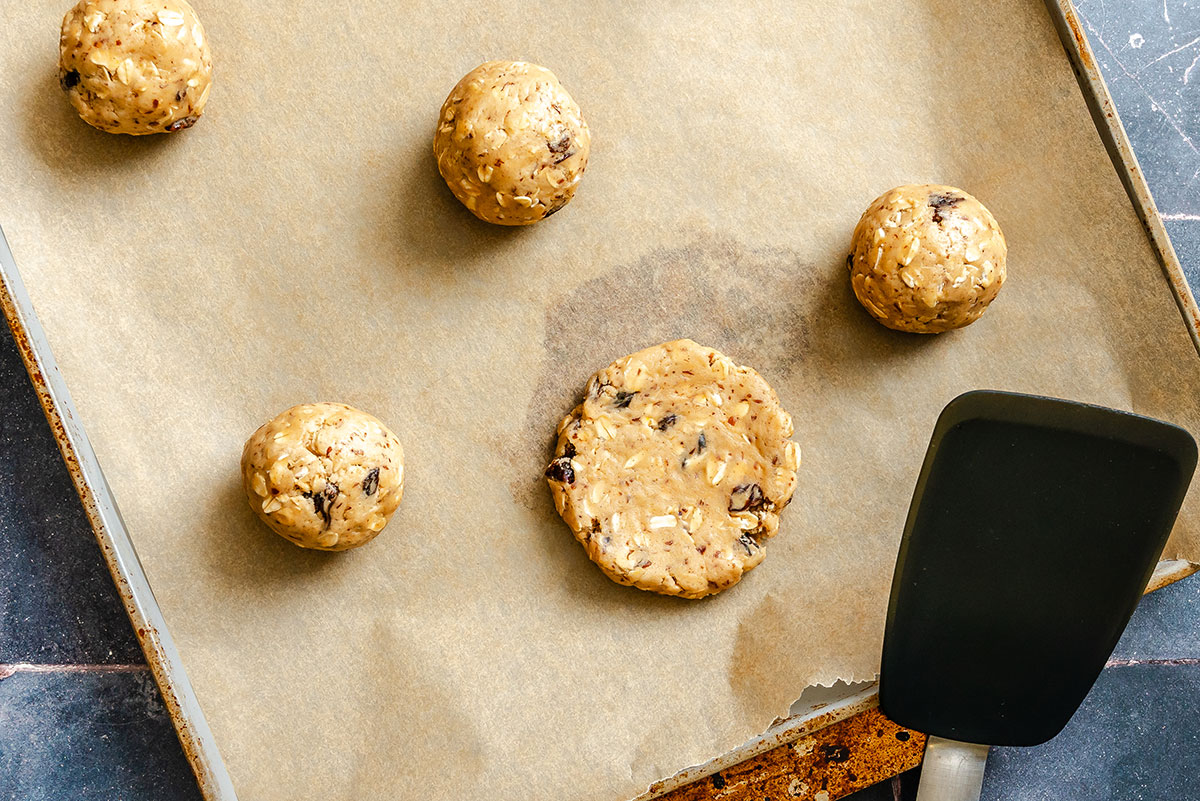 Cookie dough balls on a parchment-lined baking sheet, with one dough ball flattened by a black spatula.