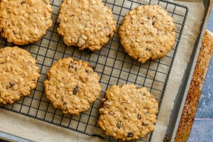 Six oatmeal cookies with raisins cooling on a black wire rack placed on a baking sheet lined with parchment paper.