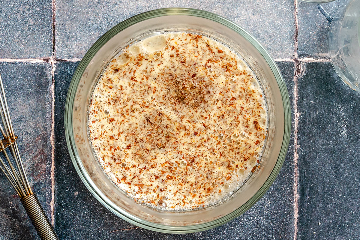 A glass bowl containing a frothy mixture with visible brown specks for vegan oatmeal cookies sits on a tiled surface next to a metal whisk.