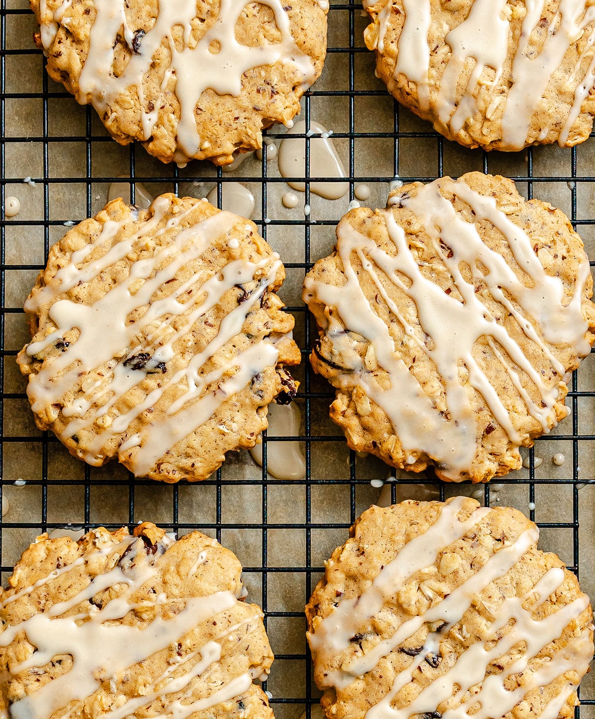 Six oatmeal cookies with white glaze drizzle cooling on a wire rack over parchment paper.