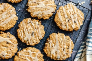 Oatmeal cookies with icing drizzle cooling on a black wire rack, with a striped towel partially visible on the side.