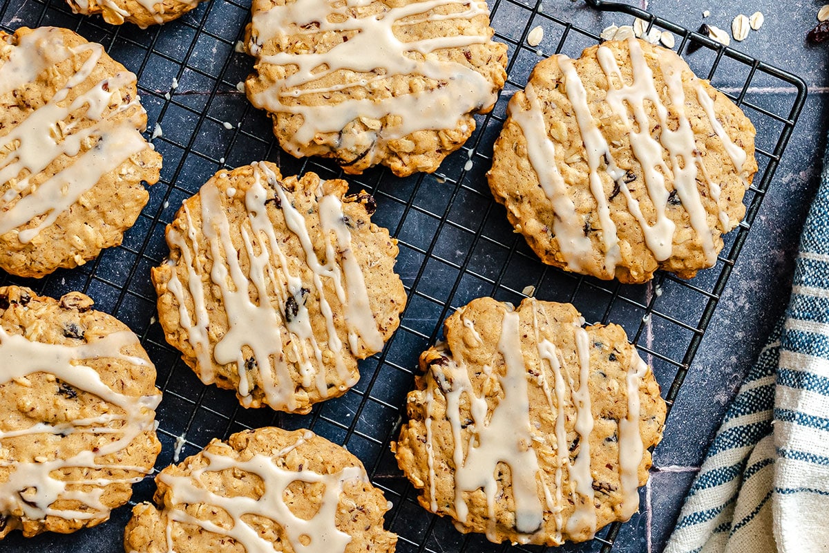 Oatmeal cookies with icing drizzle cooling on a black wire rack, with a striped towel partially visible on the side.