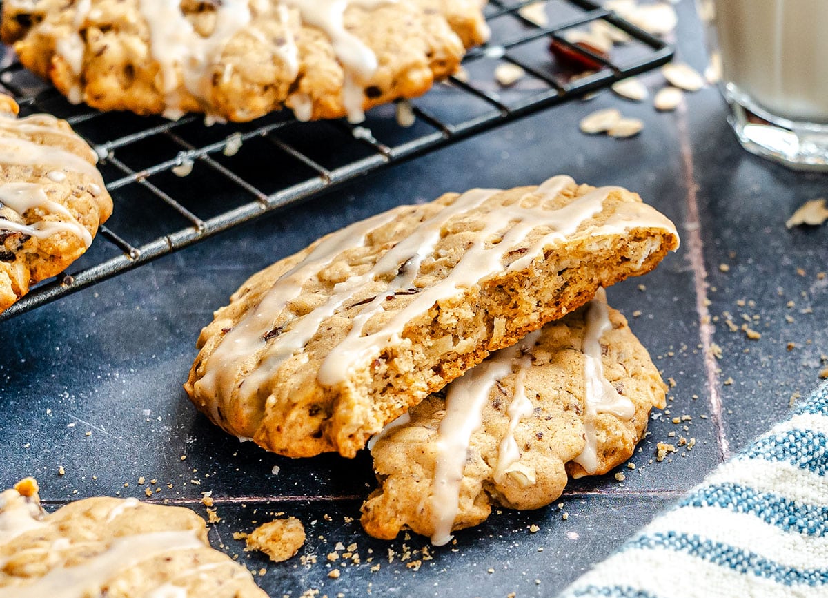An oatmeal cookie with icing is broken in half, resting on another cookie, with more cookies on a cooling rack and a glass of milk in the background.