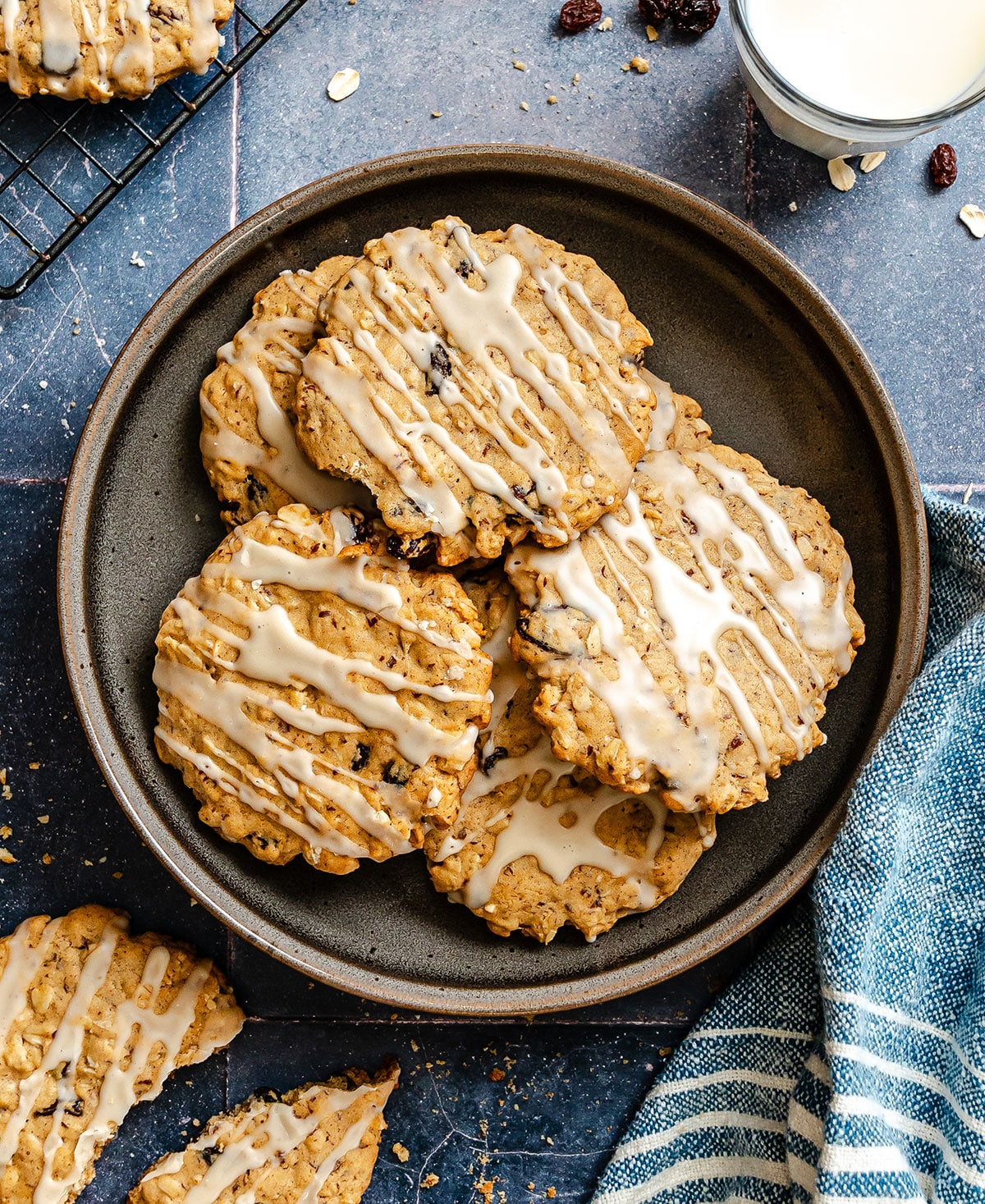 A plate of oatmeal cookies with white icing drizzle, next to a glass of milk, cooling rack, and blue striped cloth on a dark surface.