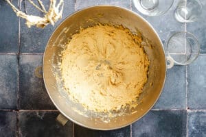 A mixing bowl filled with creamed cookie dough sits on a tiled countertop, with a mixer attachment and empty glass bowls nearby.