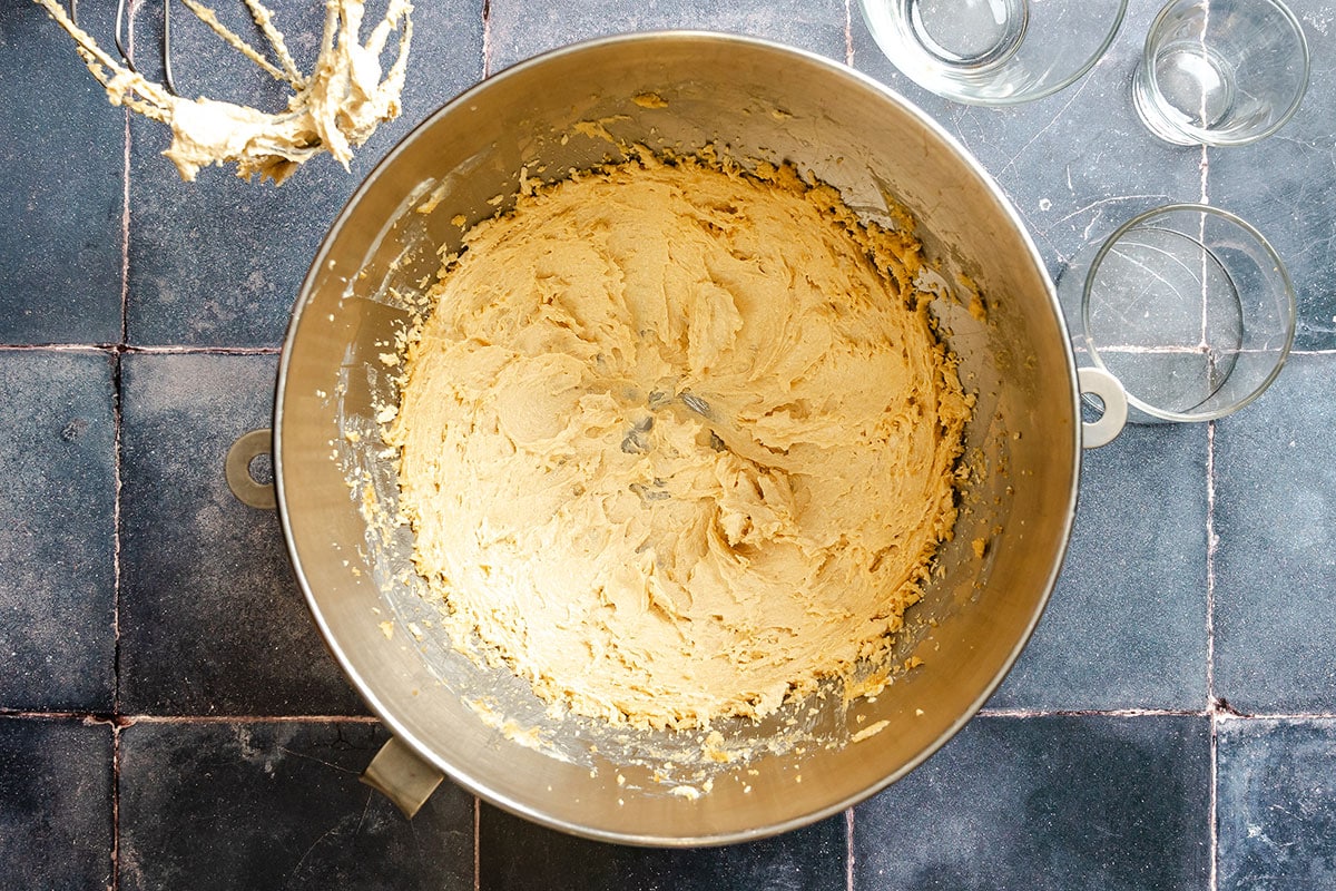 A mixing bowl filled with creamed cookie dough sits on a tiled countertop, with a mixer attachment and empty glass bowls nearby.