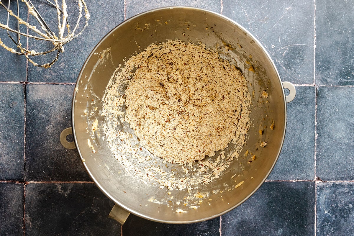 A metal mixing bowl containing cookie dough sits on a dark tiled surface, with a whisk attachment nearby.